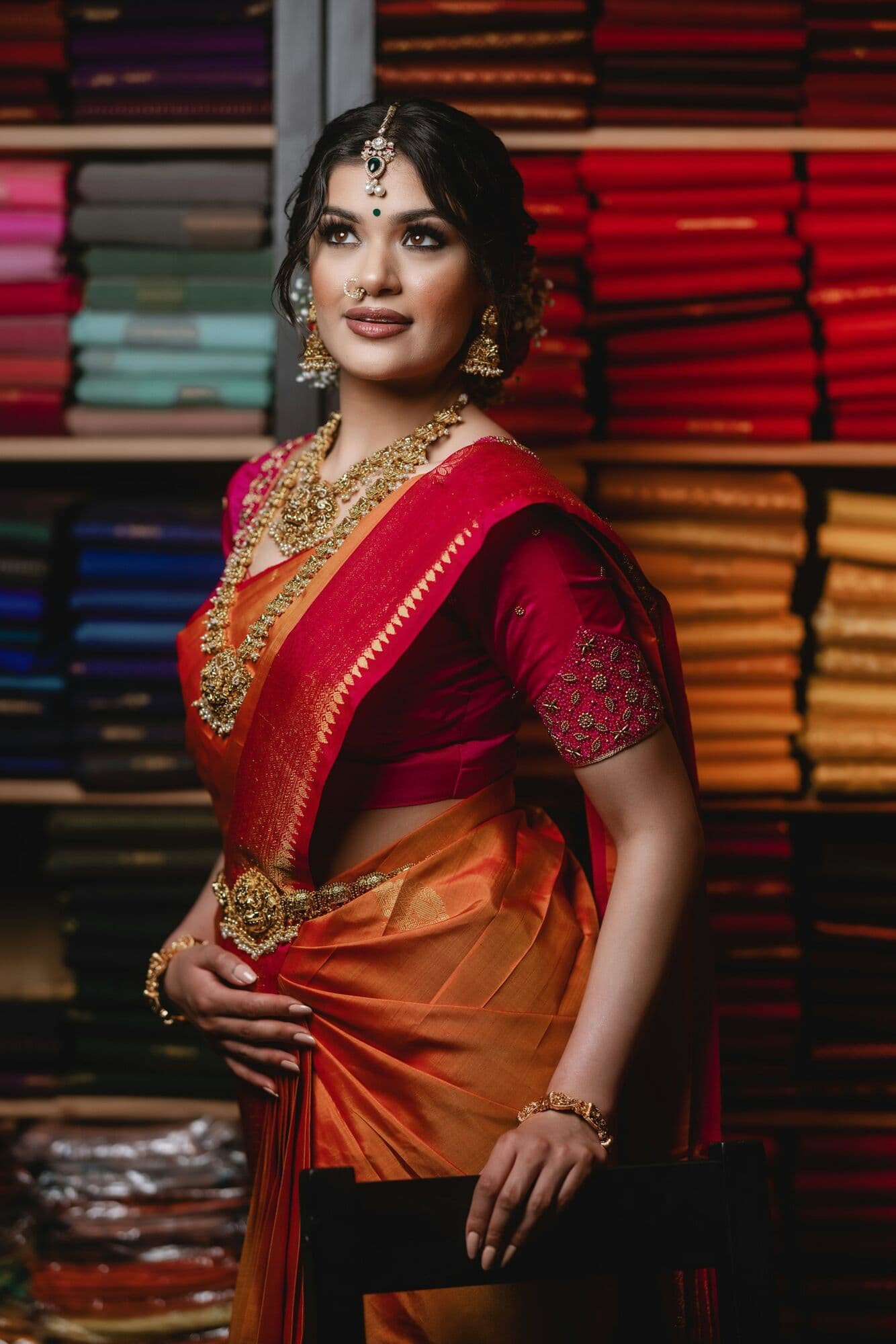 A woman in a red and orange silk saree in front of saree shelves