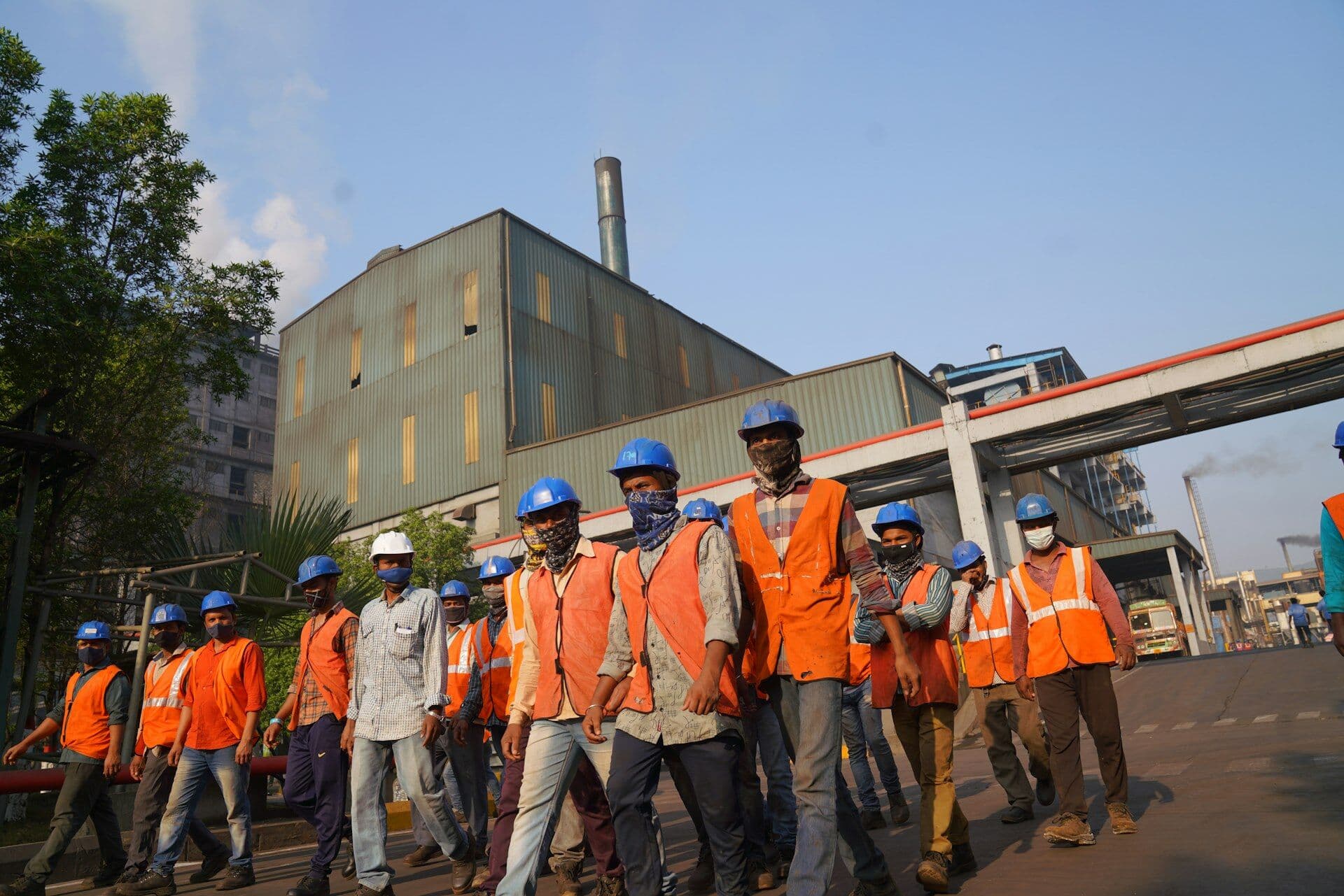 Industrial workers in orange safety vests and helmets walking past a plant